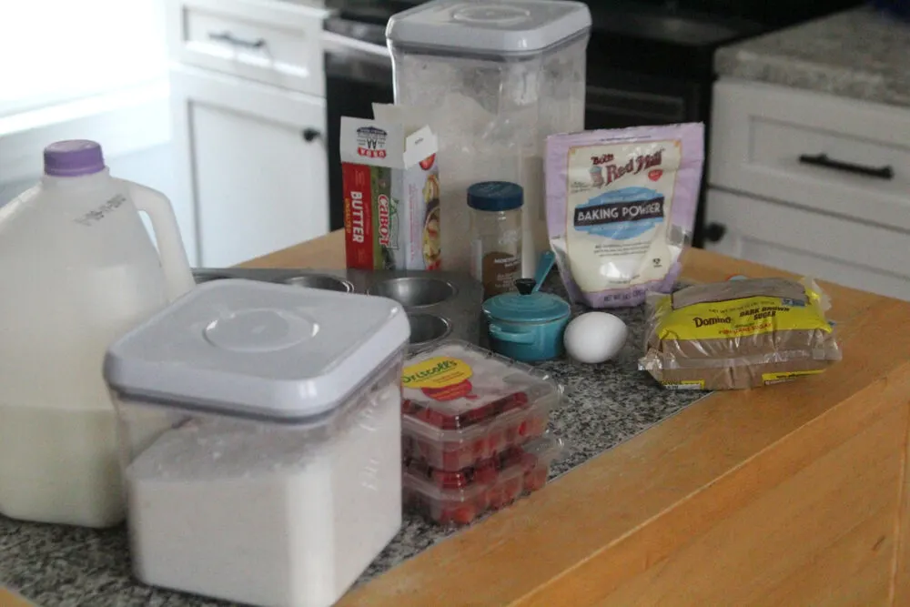Ingredients such as sugar, raspberries, milk, baking powder, an egg and butter sit on a countertop.