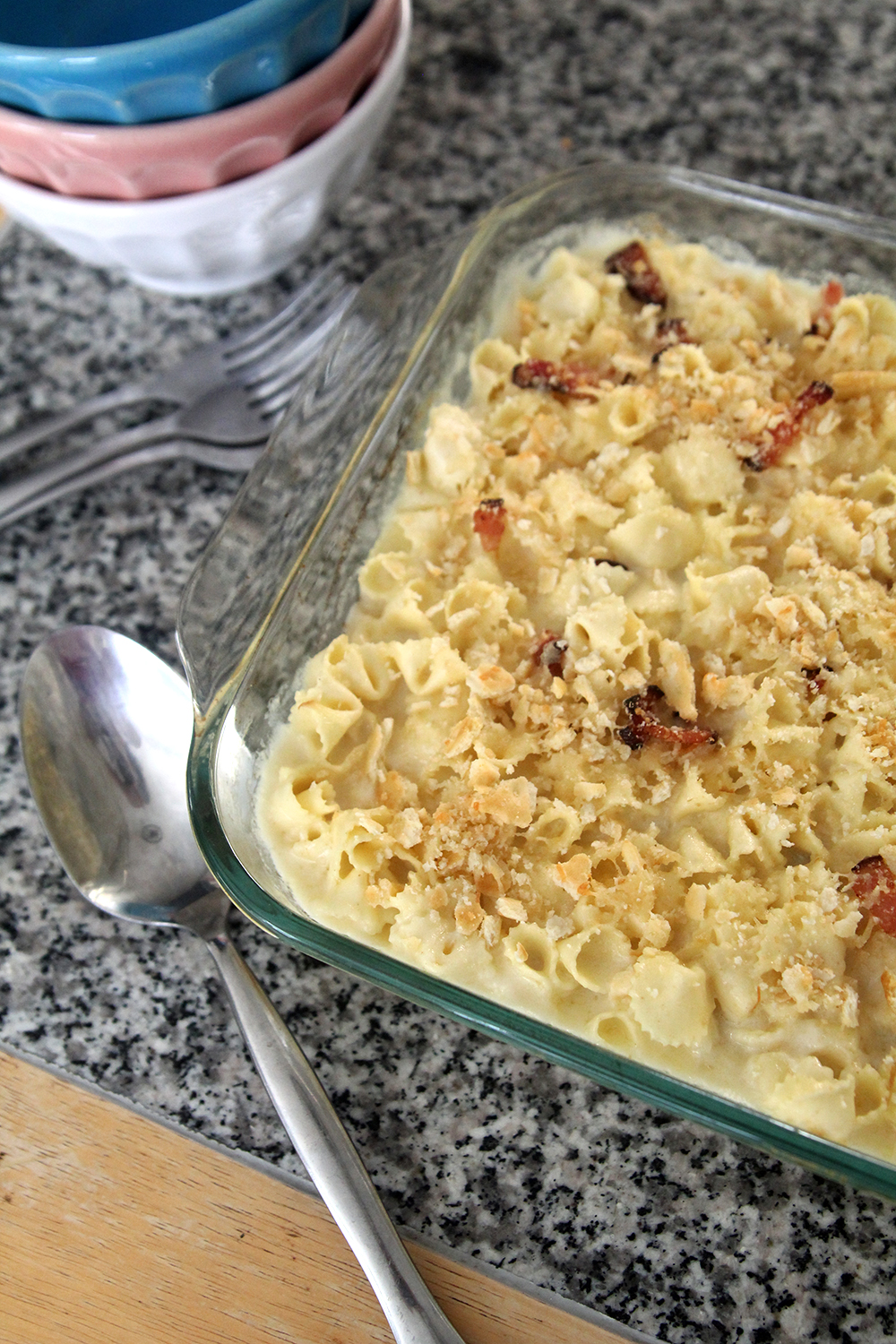 A pan of macaroni and cheese with bacon and a cracker topping is shown from above. A serving spoon sits nearby along with three forks and a stack of bowls.