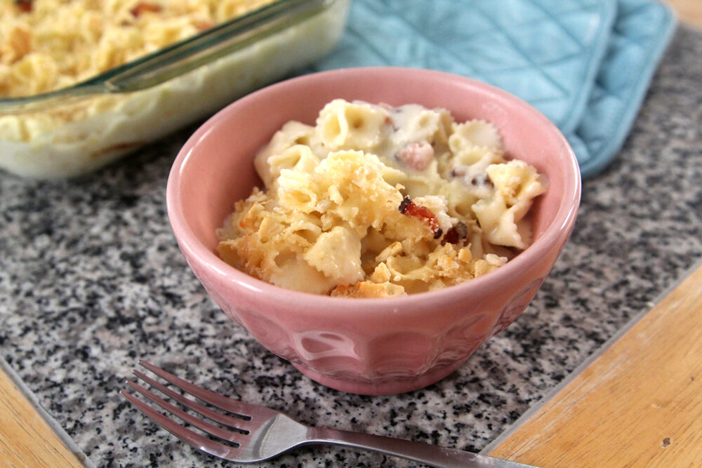 Smoked Cheddar Mac and Cheese with Bacon is portioned into a pink bowl. You can see the bacon pieces and cracker topping. Nearby is two pot holders and the pan of mac and cheese.