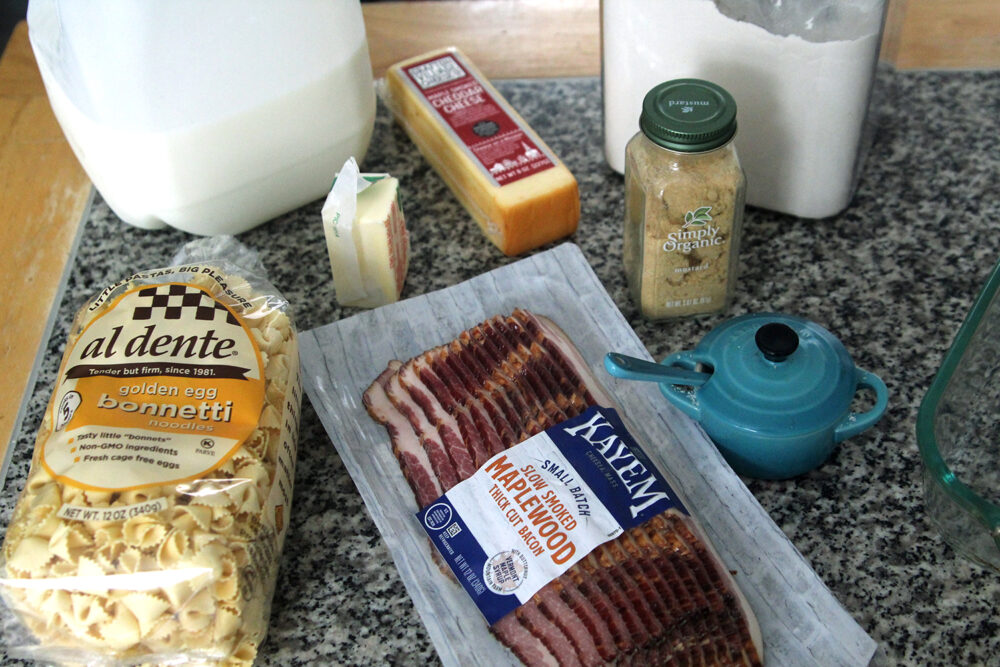 Ingredients for this macaroni and cheese recipe including pasta, milk, cheddar, bacon, mustard, butter and flour are shown.