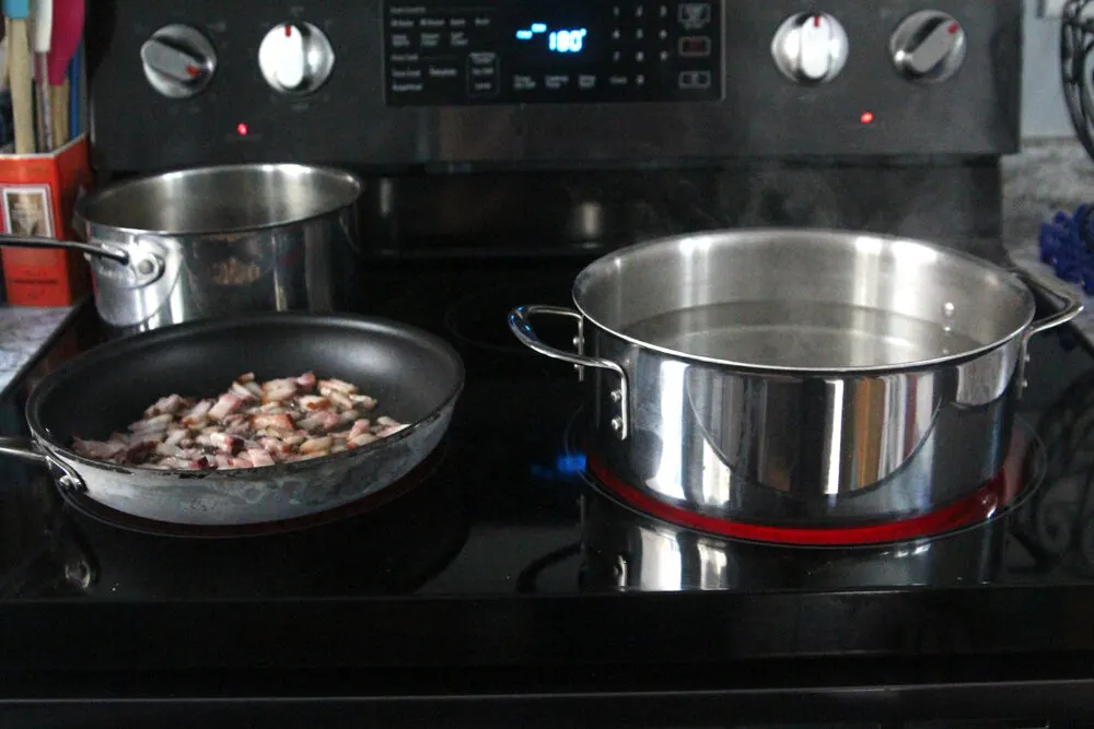 A stovetop is shown. On it is a pot of water with steam rising, a pan with bacon cooking and a saucepan that appears empty.