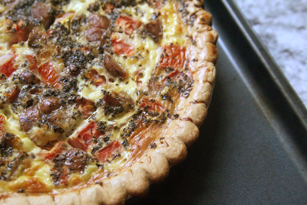 A baking sheet holds a quiche in this photo. The quiche has a pastry crust, tomatoes, sausages and other fillings. 