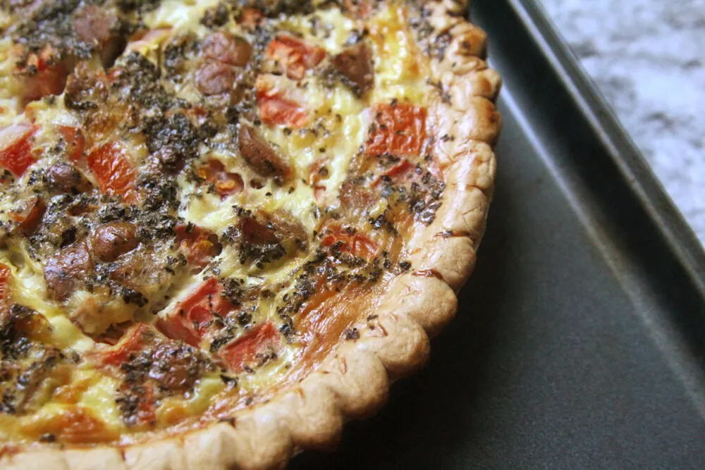 A baking sheet holds a quiche in this photo. The quiche has a pastry crust, tomatoes, sausages and other fillings.