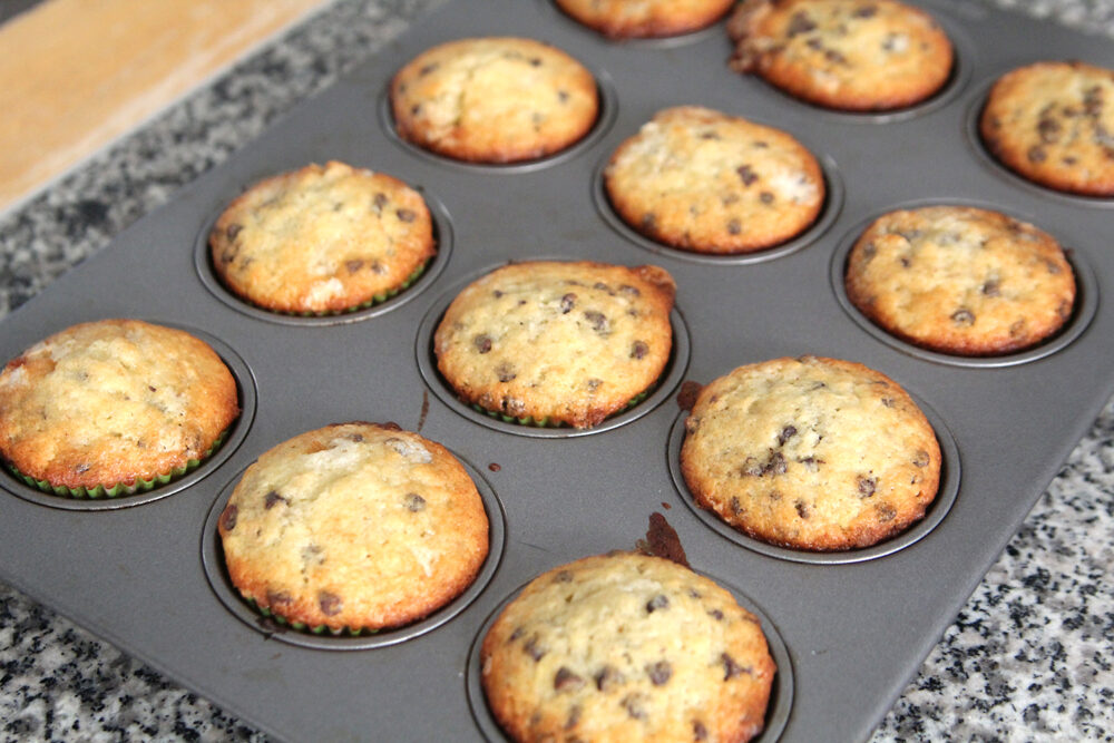 A muffin pan filled with lightly browned muffins with darker brown spots is shown on a granite countertop.