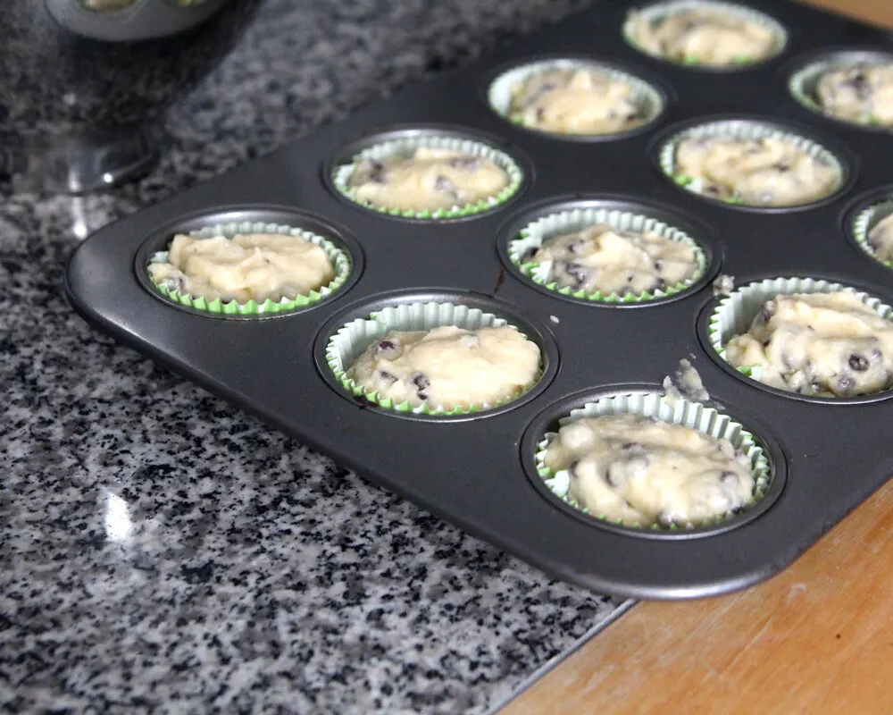 A muffin pan is shown with paper liners filled with light tan batter that has dark brown bits in it.