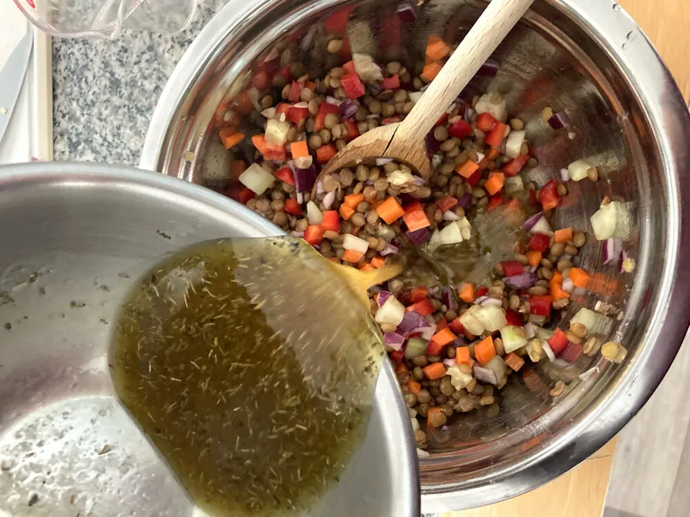 A yellow-ish vinaigrette is poured from a mixing bowl into another mixing bowl of lentils, carrots, red onions, red peppers and cucumbers. 