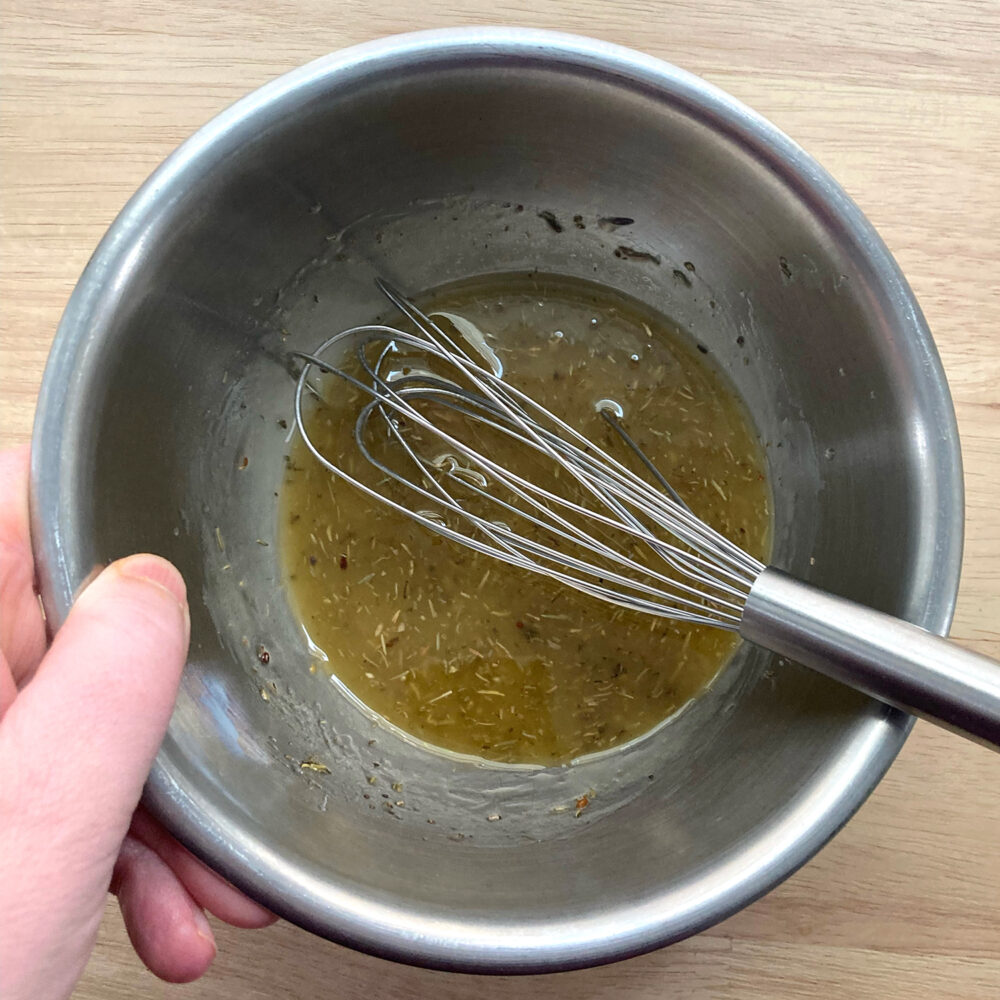 A silver mixing bowl holds a yellowish vinaigrette with herbs and a small whisk.