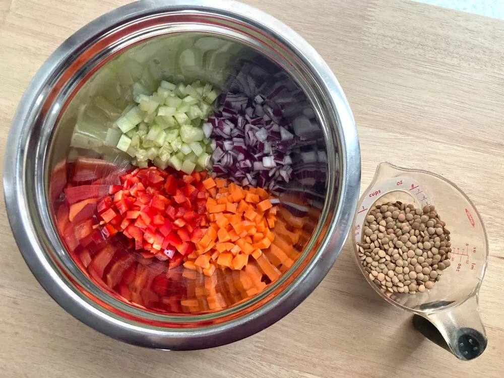 A silver mixing bowl holds red onions, cucumbers, red peppers and carrots next to a measuring cup of lentils.