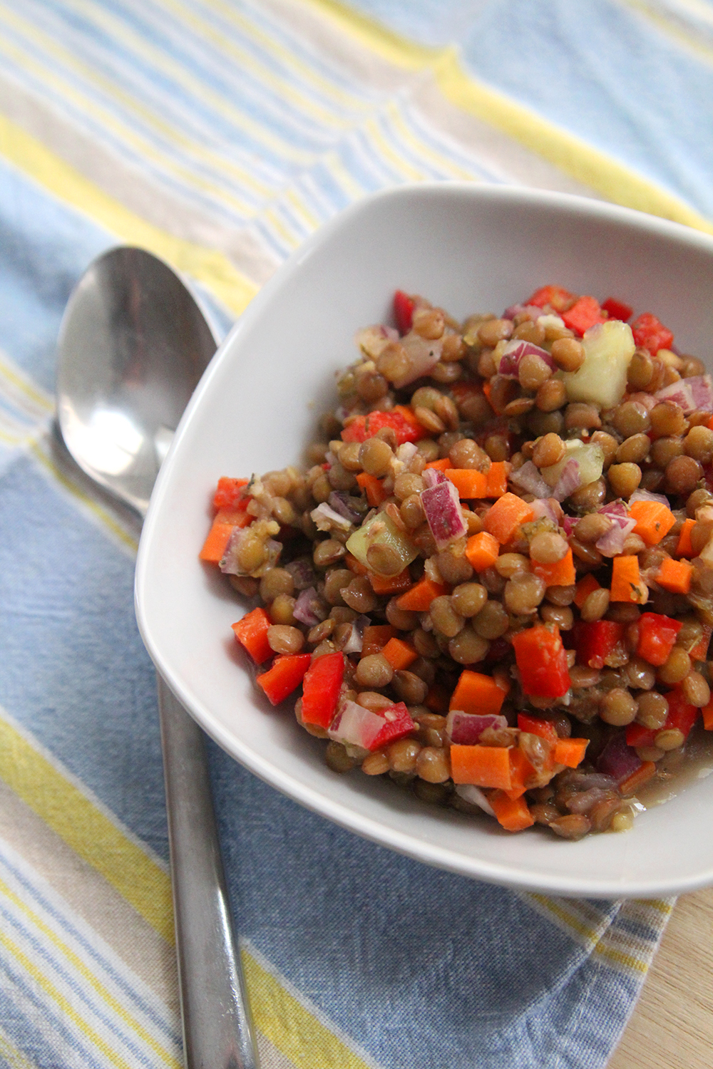 A bowl filled with cold lentil salad — lentils, red onions, cucumbers, red peppers and carrots — sits on a blue and yellow striped fabric with a spoon nearby.