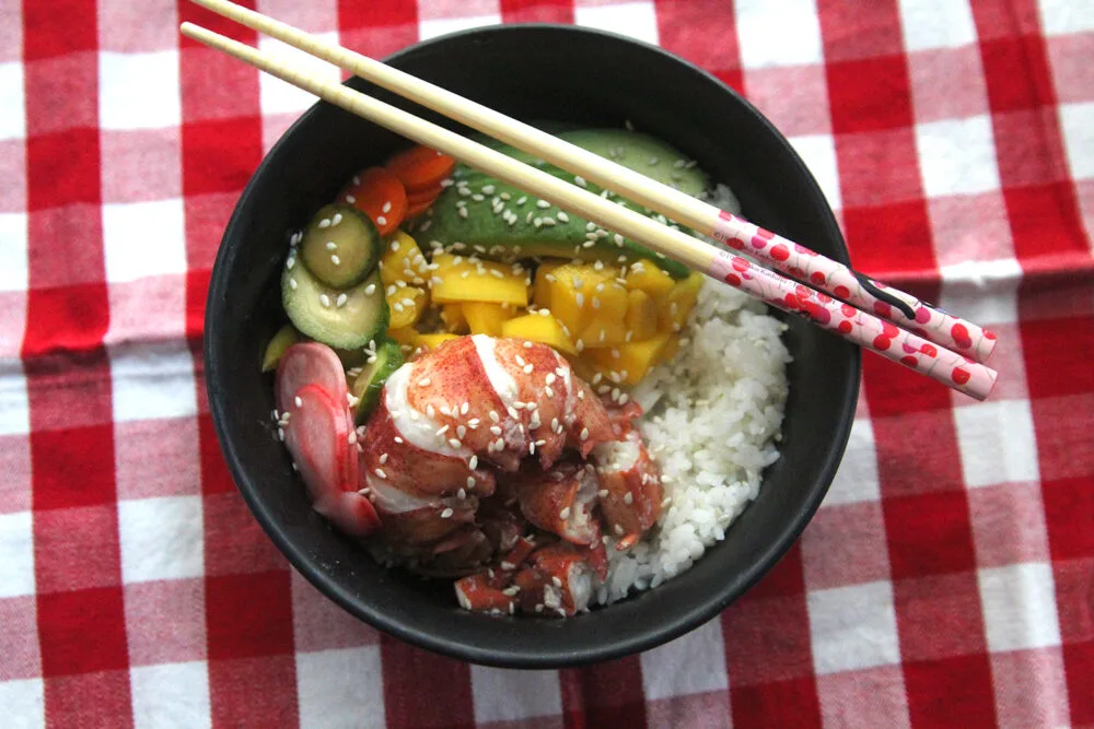 A black bowl filled with white rice, yellow mango, green avocado, succulent lobster and a trio of pickled vegetables sprinkled with sesame seeds sits on a red and white checkered cloth.