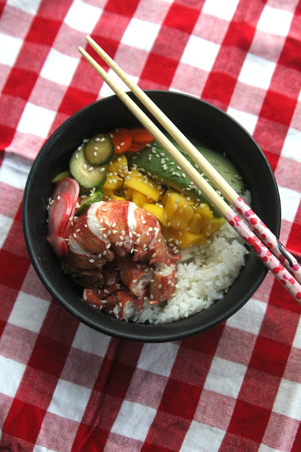 A black bowl filled with white rice, yellow mango, green avocado, succulent lobster and a trio of pickled vegetables sprinkled with sesame seeds sits on red and white checkered cloth with chopsticks perched on the bowl.