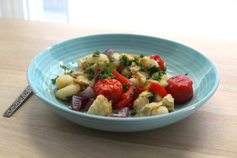 A blue bowl is shown on a wooden surface with a fork. There's golden-browned gnocchi, burst tomatoes, red onions and artichoke hearts inside. And it's all sprinkled with parsley.