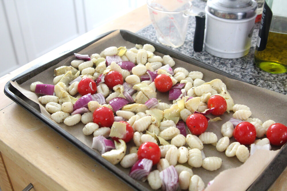 A rimmed baking sheet is shown on a counter with brown parchment paper on top. The tray is holding gnocchi, tomatoes, red onions and artichoke hearts that have been drizzled with olive oil and sprinkled with salt and pepper.