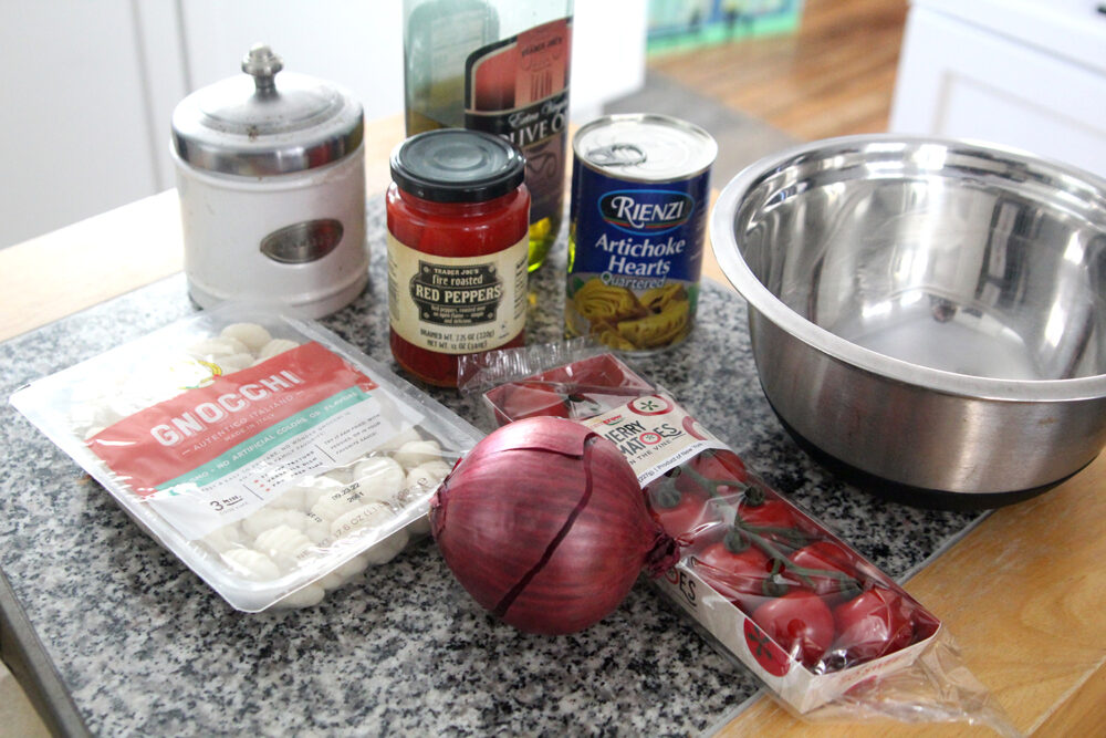 Ingredients for Sheet Pan Gnocchi are shown on a counter including gnocchi, a red onion, cherry tomatoes, roasted red peppers, artichoke hearts, olive oil and a jar with garlic. An empty mixing bowl sits nearby.