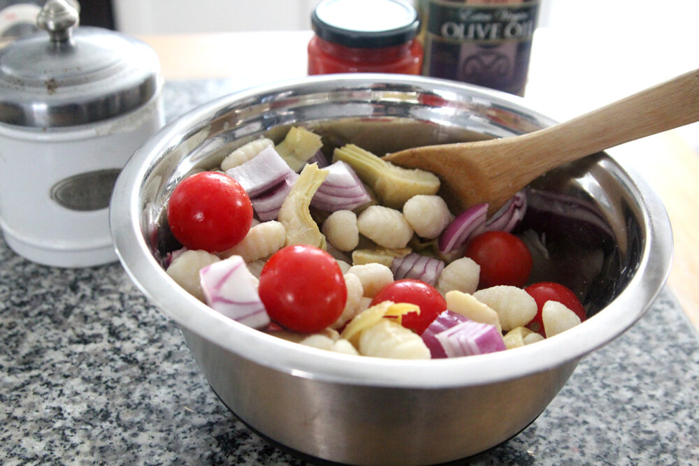 A mixing bowl is shown with red onions, gnocchi, cherry tomatoes and artichokes inside with a wooden spoon.