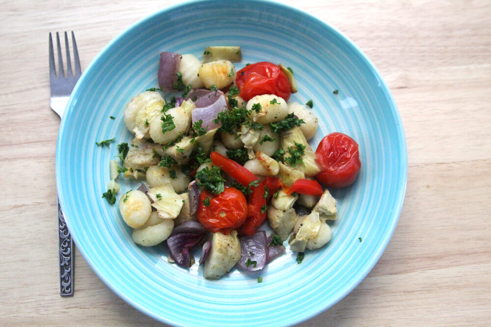 A blue bowl is shown on a wooden surface with a fork. There's golden-browned gnocchi, burst tomatoes, red onions and artichoke hearts inside. And it's all sprinkled with parsley.