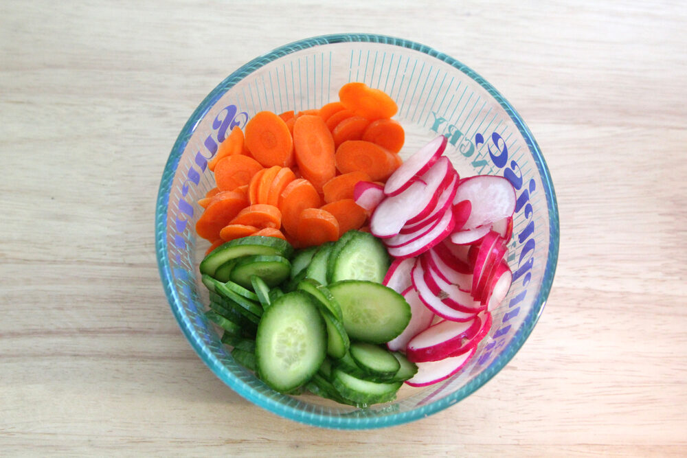 A glass bowl with writing on the sides holds orange carrot slices, red and white radish slices and green cucumber slices.