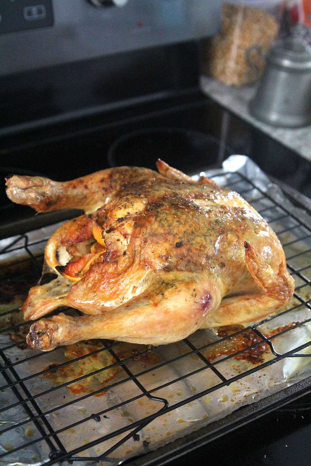 A brown and tan tinged roast chicken with orange wedges in the cavity sits on a black rack on top of a foil lined baking sheet.