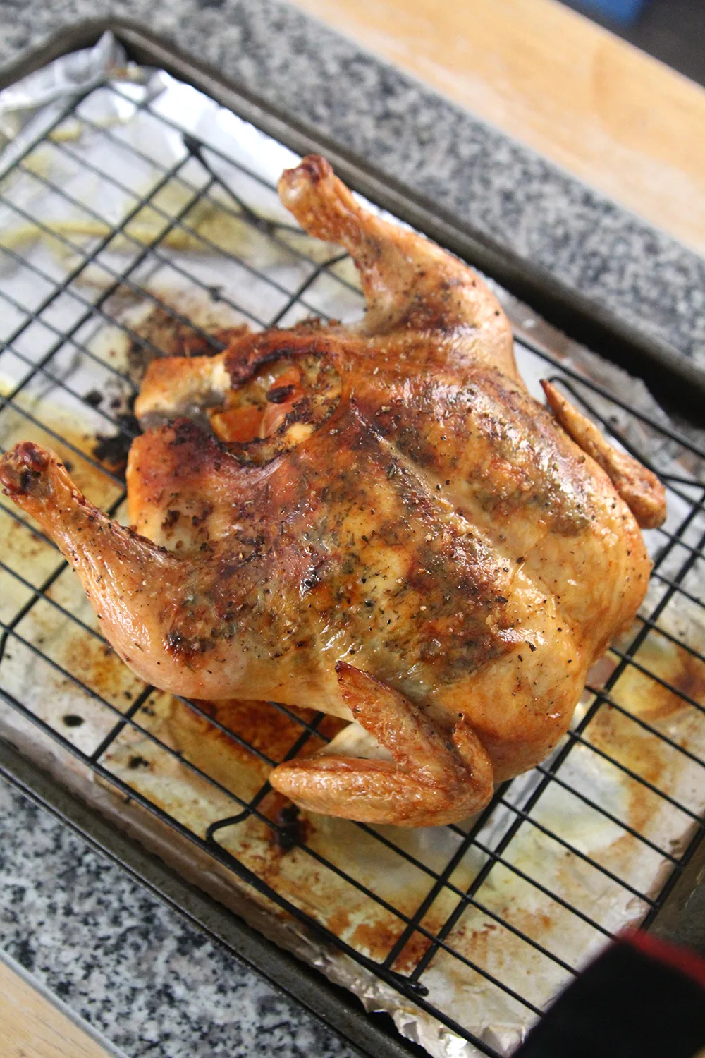 A juicy roasted chicken sits on a black rack on top of a foil-lined baking sheet. It's browned and you can see the seasonings through the skin.