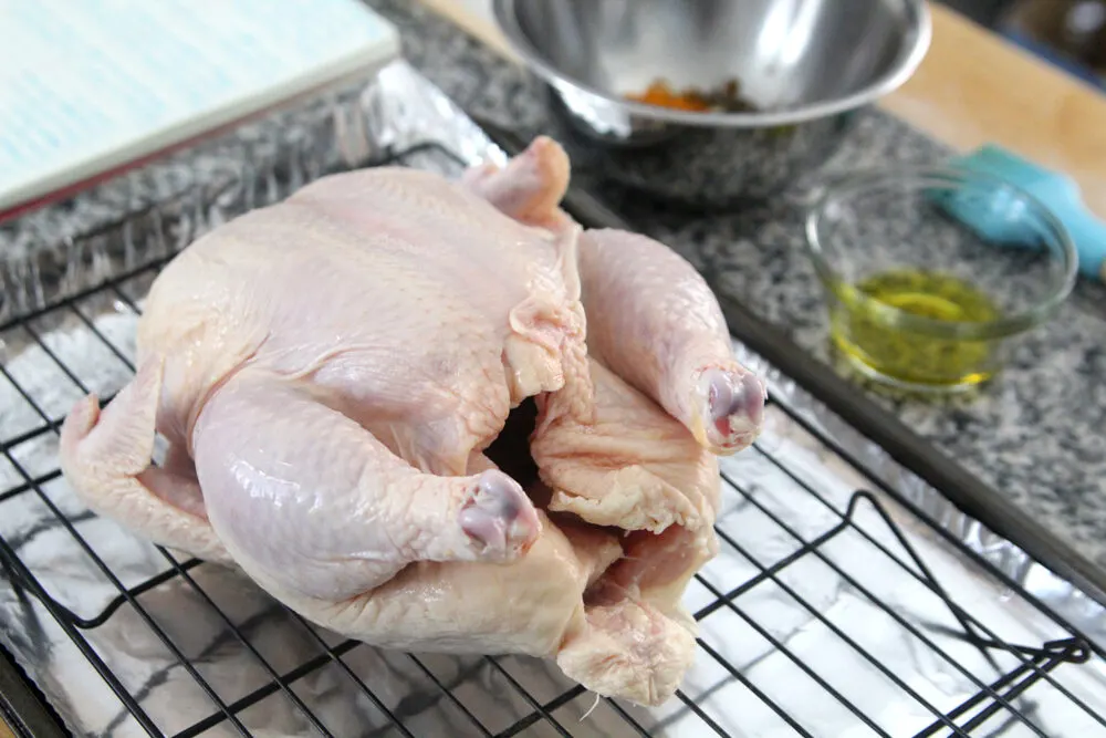 A raw whole chicken sits on a black rack on top of a foil-lined baking sheet. In the background, there is a bowl of olive oil, a bowl of seasonings and a brush.