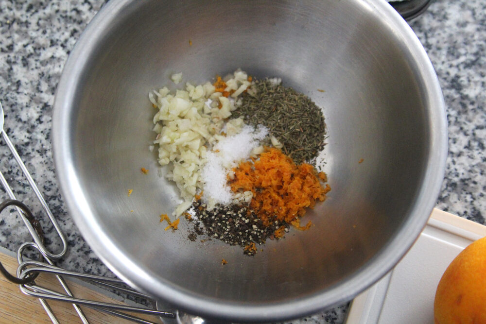 A silver bowl is filled with seasonings including minced garlic, zested orange peel, thyme, salt and pepper.