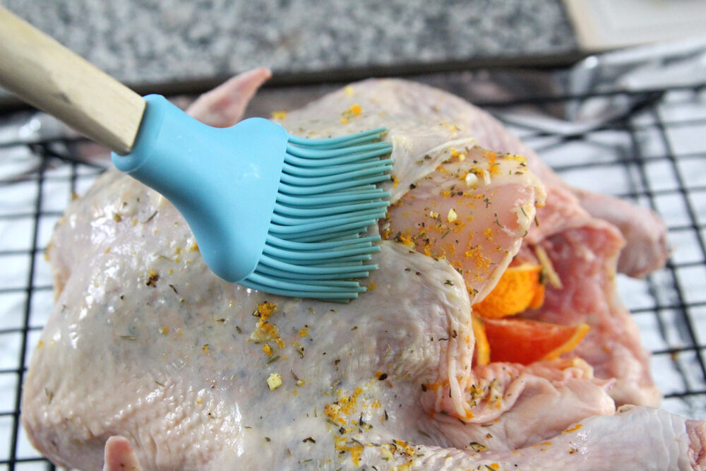 An aqua-colored brush is pressed onto the raw skin of a whole chicken. The chicken has orange wedges in the cavity and seasonings on the meat and skin.