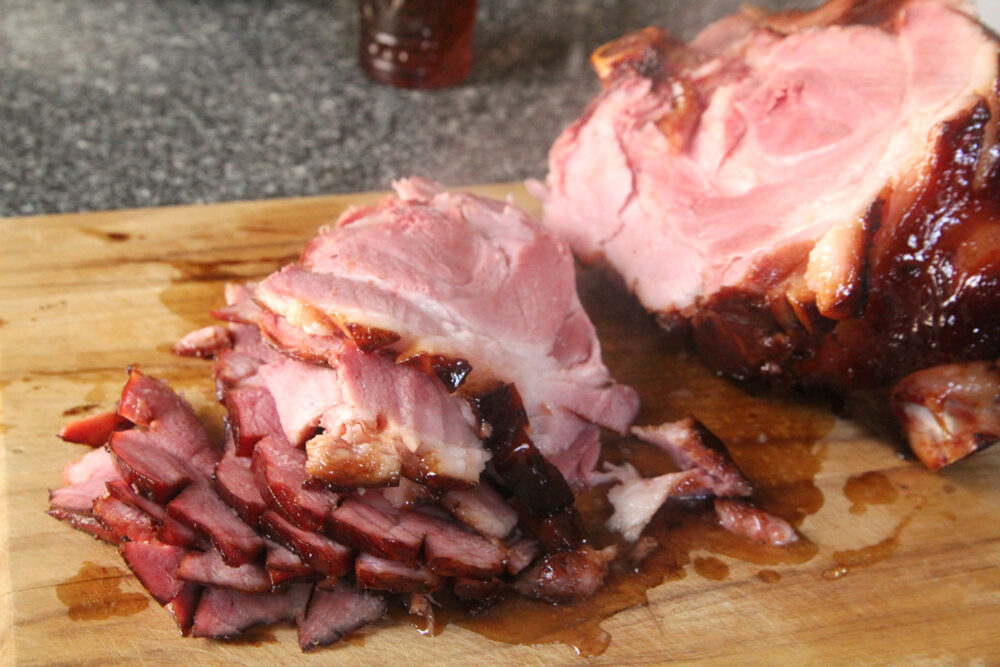 A bone-in ham has been sliced thinly and sits on a cutting board. An amber-colored glass sits in the background.