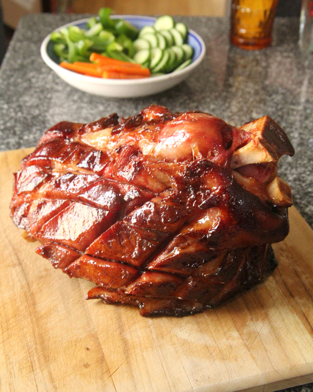 A bone-in ham that has been cut in a diamond pattern and cooked with a rich brown glaze sits on a cutting board. A bowl of cut veggies is in the background along with an amber-colored glass.