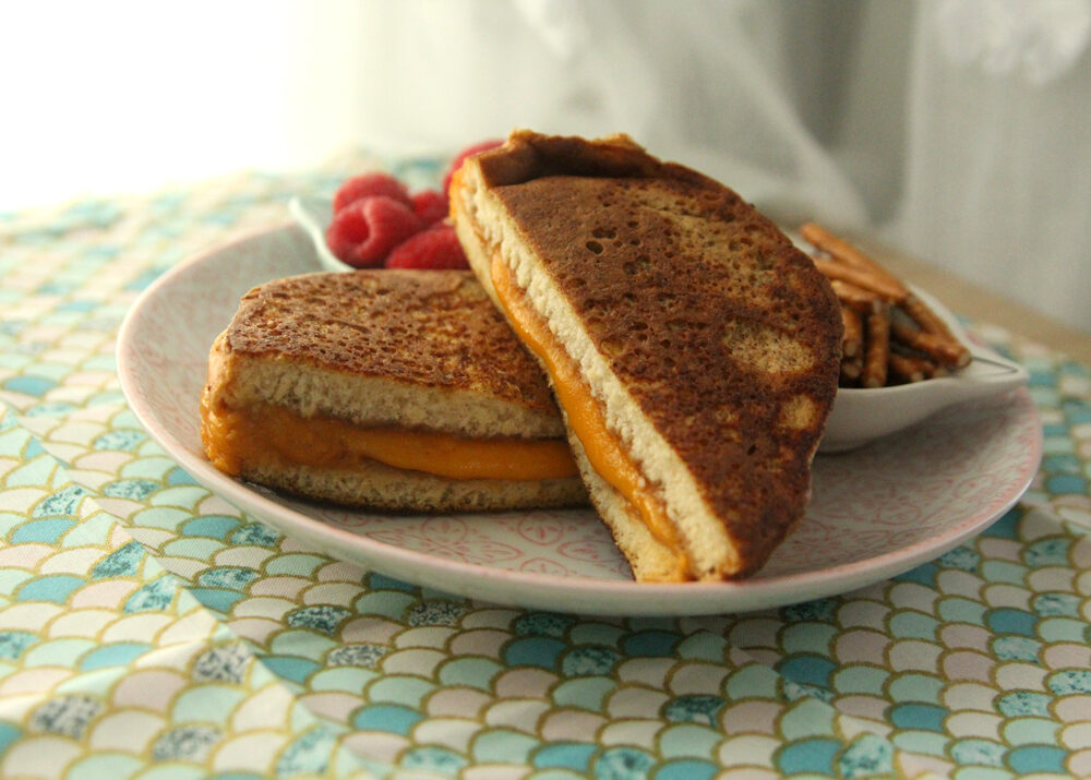 A grilled cheese sandwich with a golden brown exterior is shown on a plate with raspberries and pretzels. The sandwich is cut in half diagonally.