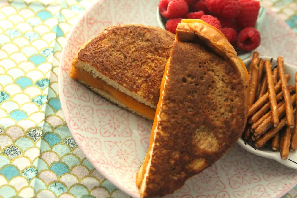 A grilled cheese sandwich with a golden brown exterior is shown on a plate with raspberries and pretzels. The sandwich is cut in half diagonally.