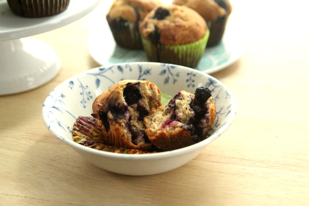 A muffin has been torn in half and sits inside a small white and blue bowl on a countertop. Nearby is a plate and a cakestand filled with Blueberry Banana Muffins. 