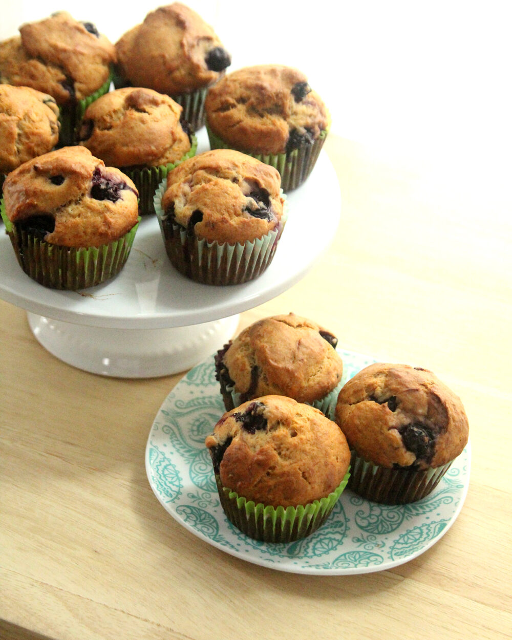A plate and a cake stand filled with Blueberry Banana Muffins sit on a counter.