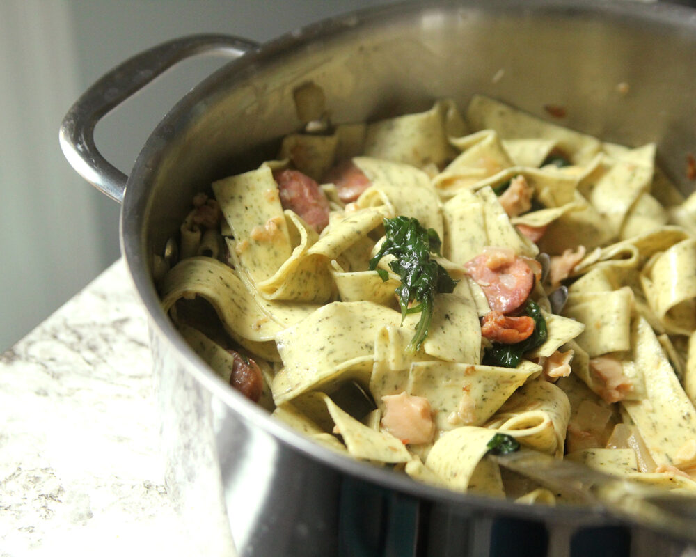 A pot of wide pasta is shown. It has bits of clam, kale and sausage intermingled in it. 