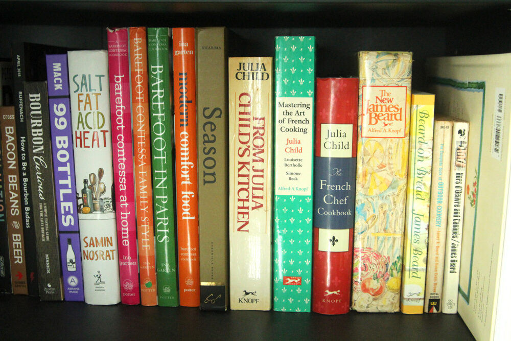 A row of cookbooks on a shelf are shown. They include books by Julia Child, James Beard, Ina Garten, Nick Sharma, Samin Nosrat and others. 