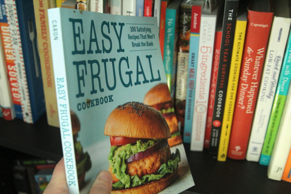 "The Easy Frugal Cookbook" by Sarah Walker Caron is held aloft with a row of cookbooks on a bookshelf behind it.