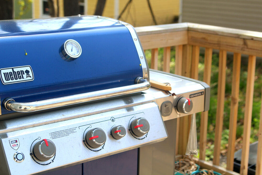 A blue and silver grill is shown on a porch. Three of the knobs are set in the on position.