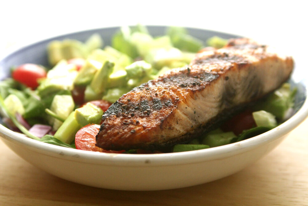A grilled piece of salmon sits on a green salad with tomatoes in a stout bowl.