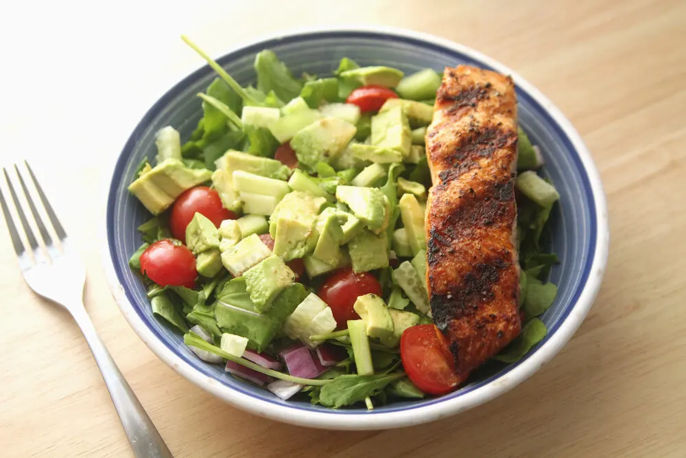 A grilled piece of salmon sits on a green salad with tomatoes in a stout bowl. A fork sits nearby.