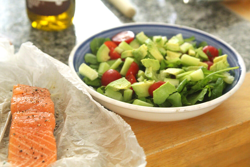 A green salad with tomatoes sits on a countertop. A salmon filet on paper sits nearby.