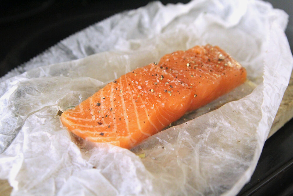 A salmon filet is shown on paper. It's seasoned with visible salt and pepper.