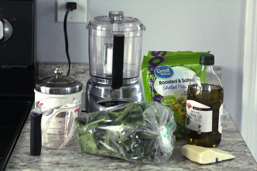 Ingredients including spinach, pistachios, olive oil and Romano cheese sit on a counter with a measuring cup and a mini food processor.