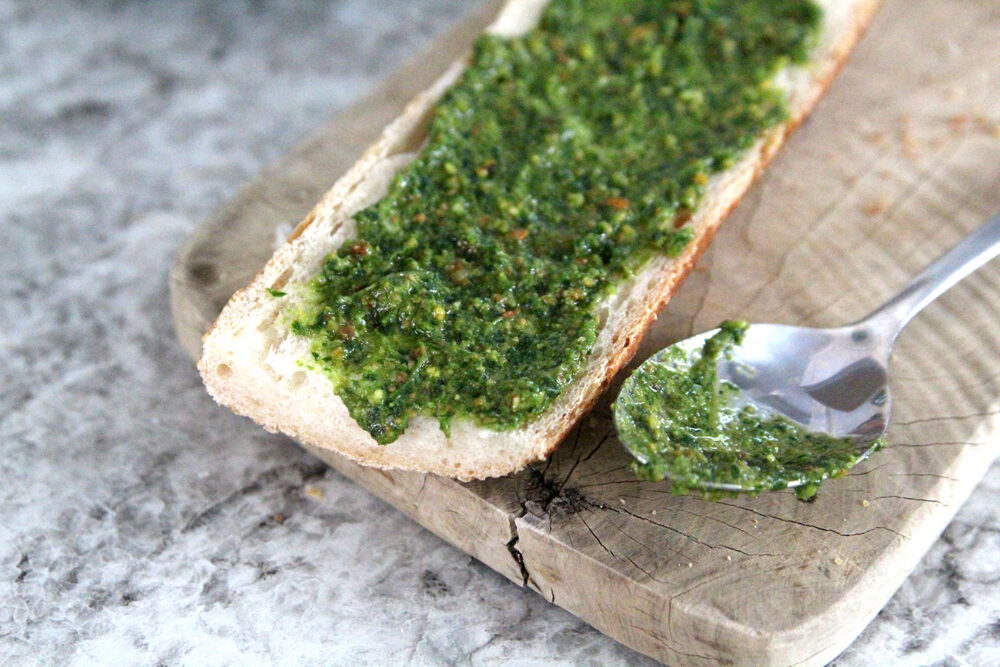 Green pesto with brown specks is spread on bread that sits on a wooden cutting board. A spoon with pesto on it sits nearby. 
