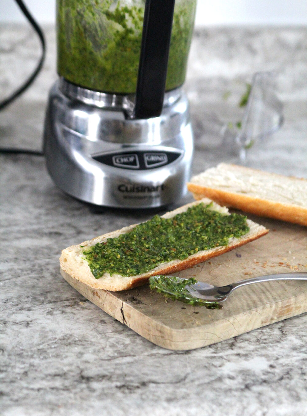Green pesto with brown specks is spread on bread that sits on a wooden cutting board. A spoon with pesto on it sits nearby. A mini food processor sits in the background.