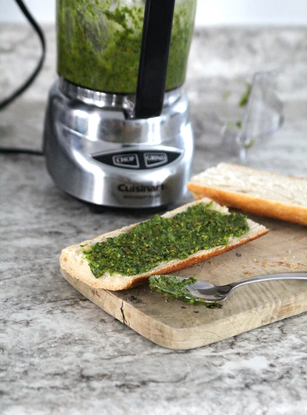 Green pesto with brown specks is spread on bread that sits on a wooden cutting board. A spoon with pesto on it sits nearby. A mini food processor sits in the background.