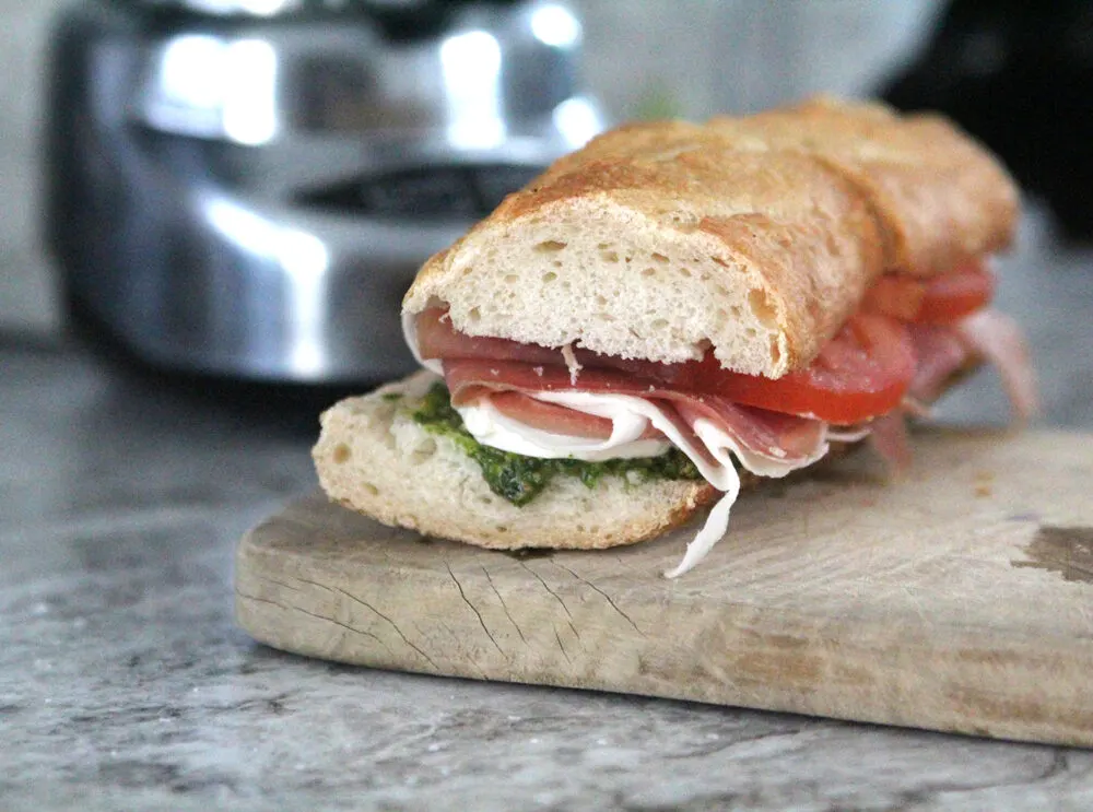 A sandwich on a French baguette sits on a wooden cutting board. Tomato, prosciutto and pesto are visible at the end.