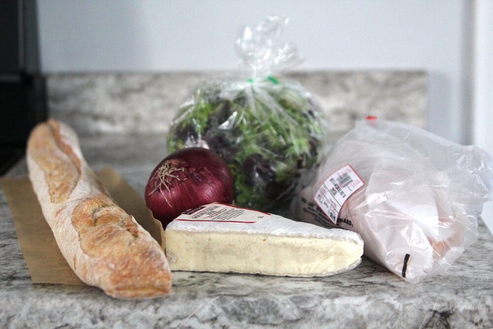 A baguette, a wedge of brie, a deli bag with turkey, a whole red onion and a bag of green and purple lettuce can be seen on a marbled countertop.
