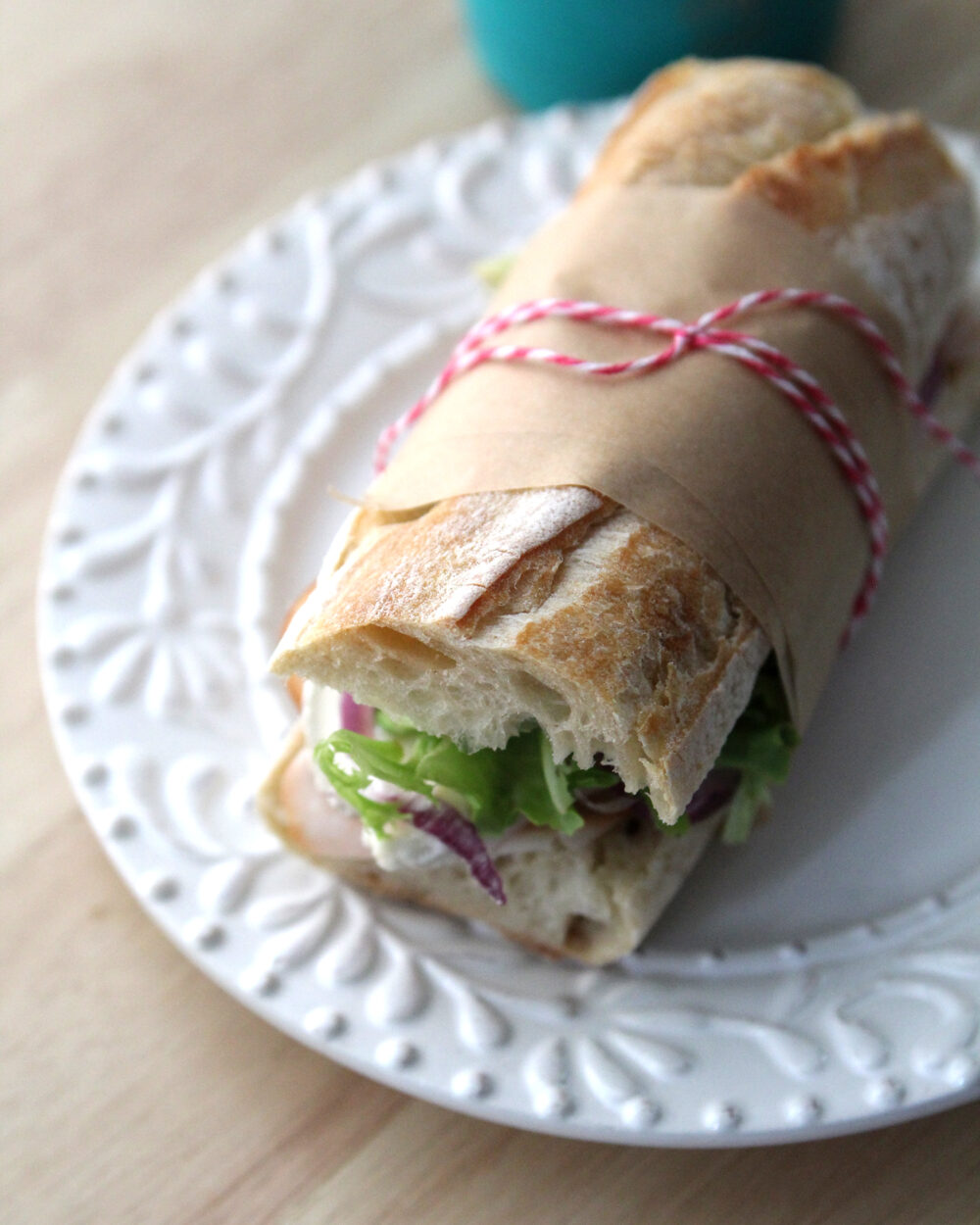 A sandwich on a baguette is shown with visible green lettuce, red onion and a bit of brie. It's sitting on a white plate and is wrapped in brown parchment paper with pink and white bakery string.