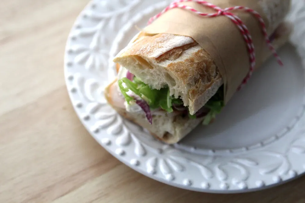 A sandwich on a baguette is shown with visible green lettuce, red onion and a bit of brie. It's sitting on a white plate and is wrapped in brown parchment paper with pink and white bakery string.