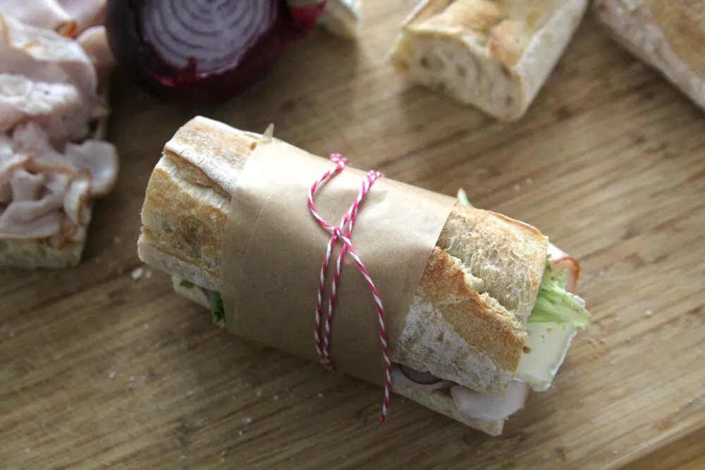 A sandwich on a baguette is shown with visible green lettuce, red onion, pinkish turkey and brie. It's sitting on a wooden cutting board and is wrapped in brown parchment paper with pink and white bakery string. Bread, red onion and turkey can be seen in the background.