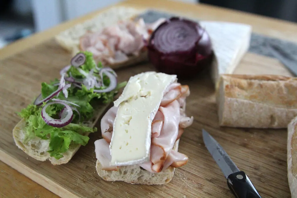 A sandwich is open on a wooden cutting board. On one half turkey is draped and topped with brie. On the other, red onion tops green lettuce. A small knife, baguette, red onion, turkey and brie can be seen in the background.